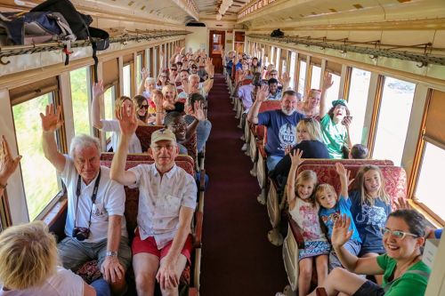 A lively group of passengers on a train, all smiling and waving from their seats.