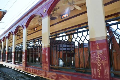 A vintage train interior featuring wooden benches and decorative arches with fans and lights.