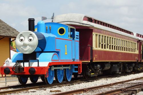 A blue train with a smiling face next to a vintage red passenger car on a sunny day.