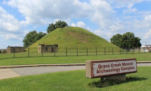 Grave Creek Mound, a large grassy burial mound, with a sign indicating the Archaeology Complex in a park setting.