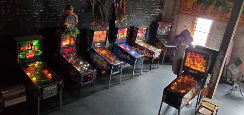A row of colorful pinball machines in a dimly lit room with vintage decor and a mannequin figure.
