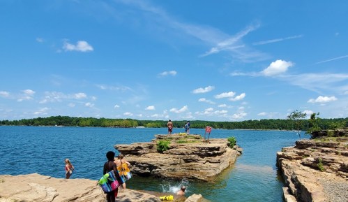 People enjoying a sunny day by the water, jumping off rocks into a lake surrounded by trees and blue skies.