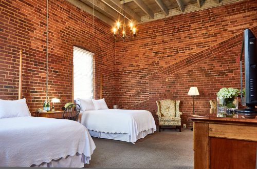 Cozy bedroom with two beds, exposed brick walls, a vintage chair, and a TV, featuring warm lighting and a welcoming atmosphere.