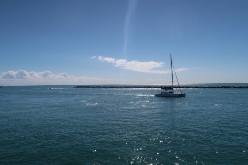 A sailboat glides across calm blue waters under a clear sky, with a rocky jetty in the background.