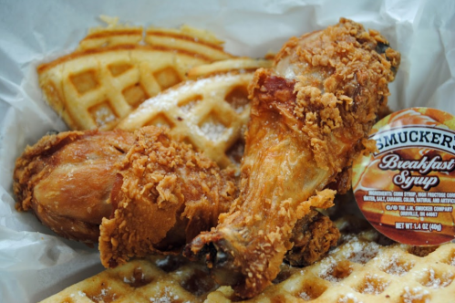 A serving of fried chicken legs with waffles and a packet of Smucker's breakfast syrup on a white paper tray.