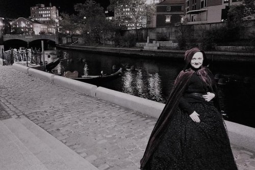 A woman in a dark cloak stands by a cobblestone path beside a river at night, with boats and city lights in the background.
