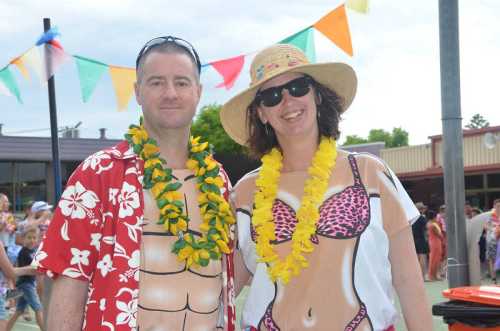 A man and woman in festive attire with leis, smiling at a lively outdoor event decorated with colorful bunting.