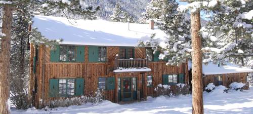 A rustic wooden house with green shutters, surrounded by snow-covered trees and mountains in a winter landscape.