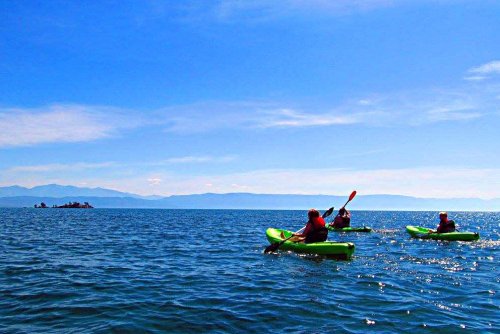 Three kayakers paddle on a calm lake under a clear blue sky, with distant mountains visible on the horizon.