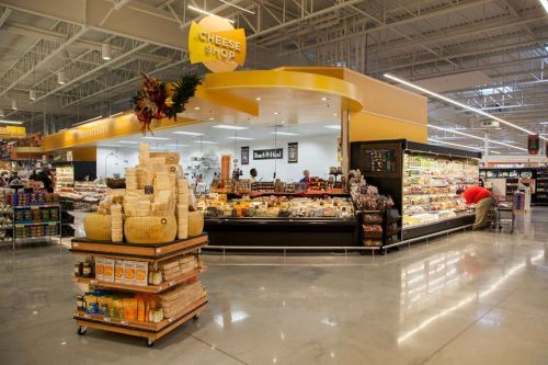 A cheese shop in a grocery store, featuring a display of various cheeses and a shopper browsing nearby.