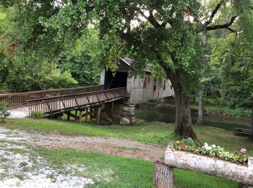 A rustic covered bridge spans a small river, surrounded by lush greenery and a gravel path.