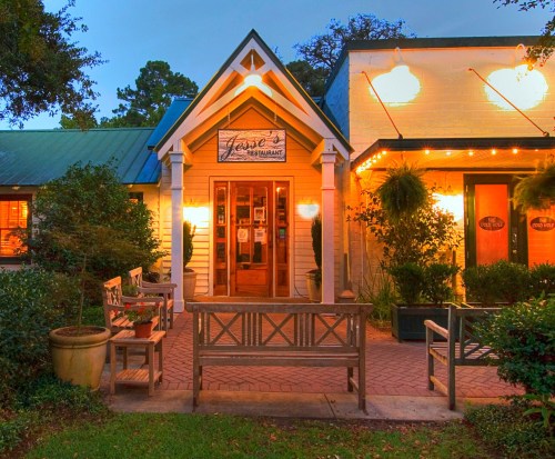 Charming restaurant exterior with warm lighting, benches, and lush greenery, inviting guests to enter.