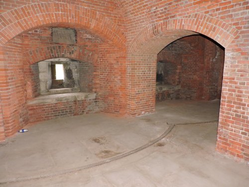 Interior of a brick structure with arched openings and a stone floor, featuring two alcoves and a faint track on the ground.