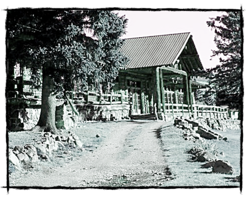 A rustic building surrounded by trees, with a gravel path leading to its entrance. The image has a vintage black-and-white effect.