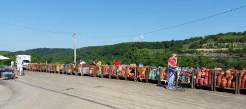 A market scene with people browsing colorful goods displayed on tables along a rural road, surrounded by green hills.