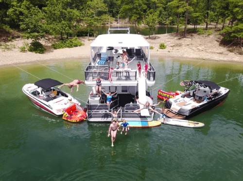 Aerial view of a large boat with people swimming, lounging, and enjoying water activities in a scenic lake setting.