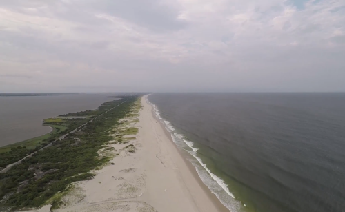 Aerial view of a sandy beach stretching along the coastline, with calm waves and greenery nearby under a cloudy sky.