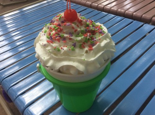 A colorful ice cream sundae in a green bucket, topped with whipped cream, sprinkles, and a cherry.