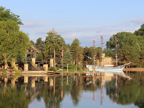 A serene lakeside scene featuring a wooden ship docked near a stone building and lush greenery, reflecting in the water.