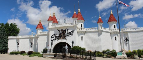 A whimsical castle-like building with red turrets and white walls, surrounded by greenery and a blue sky.