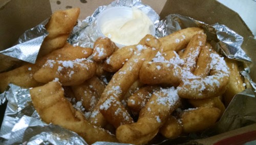 A box of golden, fried dough twists dusted with powdered sugar, accompanied by a small cup of dipping sauce.