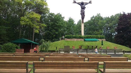 A large statue of Christ stands on a hill, overlooking empty wooden benches and a small pavilion in a green park.