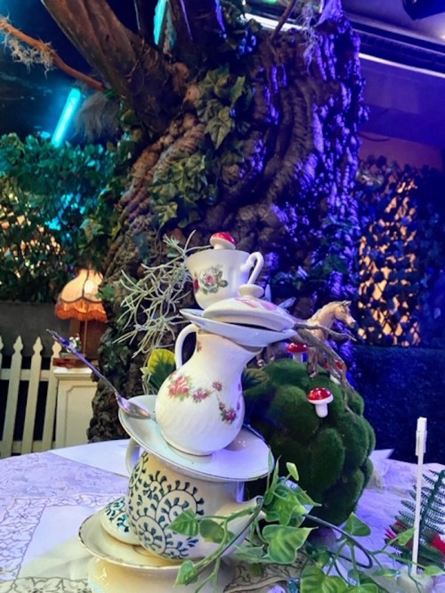 A whimsical display of stacked teapots and cups surrounded by greenery and a tree, creating a magical atmosphere.