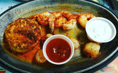 A sizzling platter with seasoned shrimp, crab cake, and dipping sauces, served with sliced potatoes.