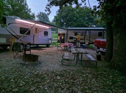 Two campers parked under trees, with a picnic table and fire pit in a gravel area, illuminated by soft lights.