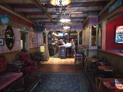 Cozy pub interior with wooden beams, vintage decor, and a bar in the background. Warm lighting and seating areas visible.