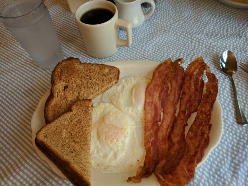 A plate with two fried eggs, crispy bacon strips, and two slices of toasted bread, with coffee cups in the background.