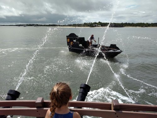 A child watches as a boat sprays water while navigating through a calm body of water.