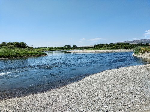 A serene river flows through a rocky shore, surrounded by lush greenery under a clear blue sky.