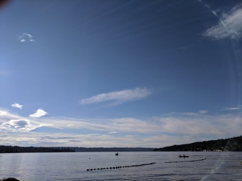 A serene lake scene under a clear blue sky with a few clouds, featuring boats on the water and trees in the background.