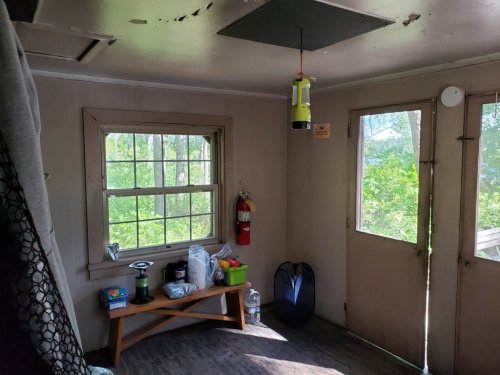 A small, sunlit room with a table, window, and door, featuring a fire extinguisher and a hanging lantern.