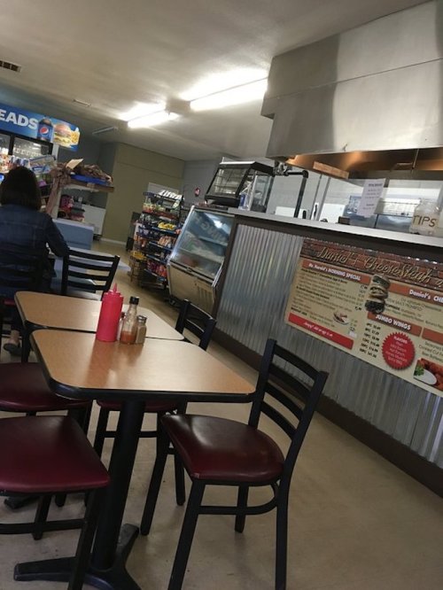 Interior of a small diner with tables, a counter, and a menu board. Shelves stocked with snacks in the background.