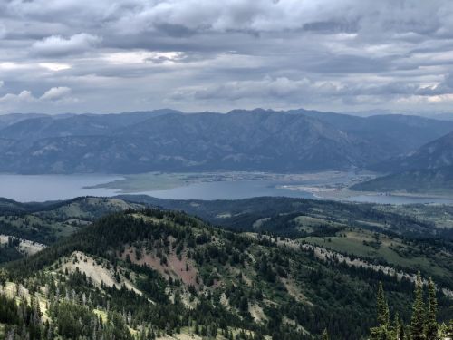 A panoramic view of mountains and a lake under a cloudy sky, with lush green hills in the foreground.