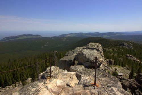 A rocky summit overlooks a vast green landscape of mountains and forests under a clear blue sky.