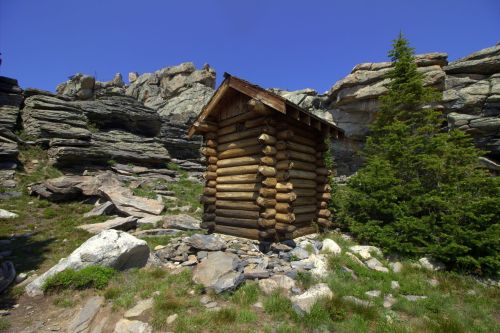 A rustic log cabin surrounded by rocky terrain and greenery under a clear blue sky.