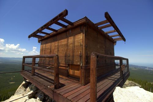 A wooden lookout tower perched on a rocky outcrop, surrounded by lush green forest and a clear blue sky.