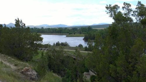 A serene river flows through a lush landscape, surrounded by trees and distant mountains under a partly cloudy sky.