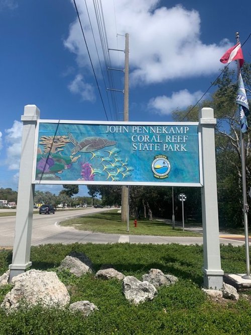 Sign for John Pennekamp Coral Reef State Park, featuring colorful marine life and flags, set against a blue sky.