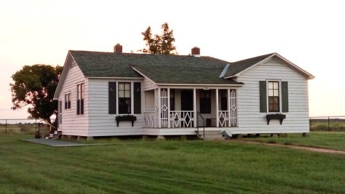 A white, single-story house with a green roof, surrounded by grass and trees, at sunset.