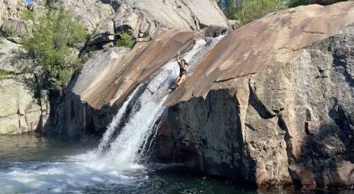 A person slides down a rocky waterfall into a clear pool surrounded by boulders and greenery.