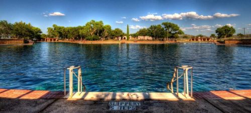A serene swimming area with clear water, steps leading in, and lush greenery under a blue sky with fluffy clouds.