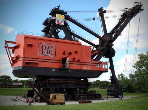 A large orange and black dragline excavator on display outdoors, with a blue sky and clouds in the background.