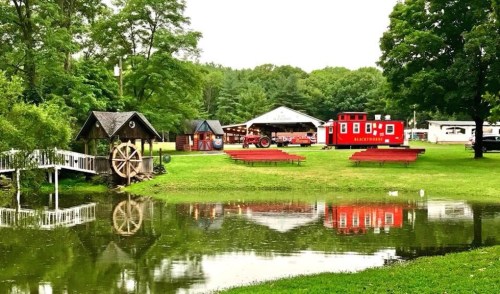 A serene park scene featuring a pond, a water wheel, and a red building with picnic tables under green trees.