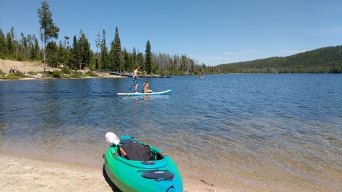 A serene lake scene with kayakers and paddleboarders, surrounded by trees and a sandy beach.