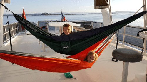 Two children play in colorful hammocks on a boat deck, with a scenic view of water and hills in the background.