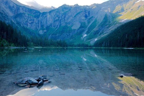 A serene mountain lake reflecting surrounding peaks and trees, with clear water and rocky foreground.